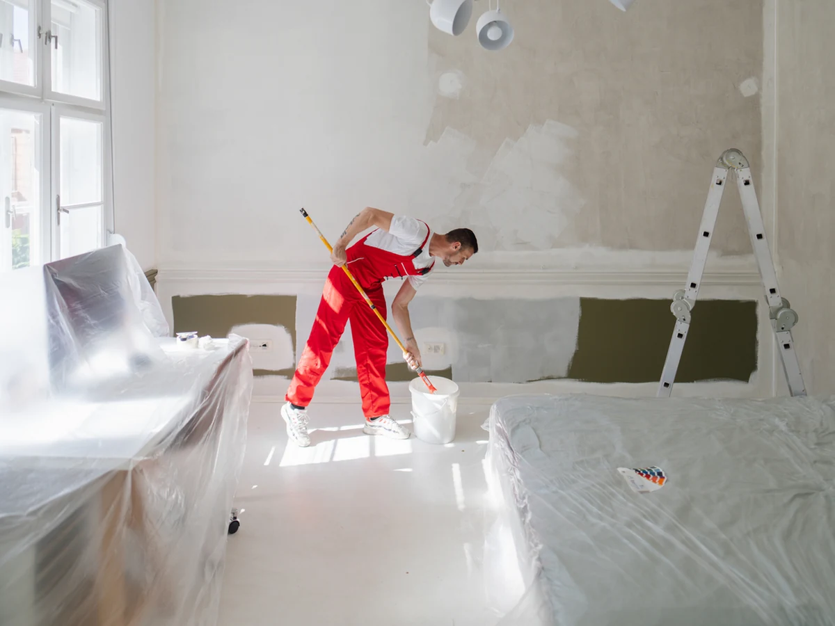 painting-decorating-painter-red-overalls-bradford-decorators A painter in red overalls rolling a wall in a room covered in dust sheets, with a stepladder behind